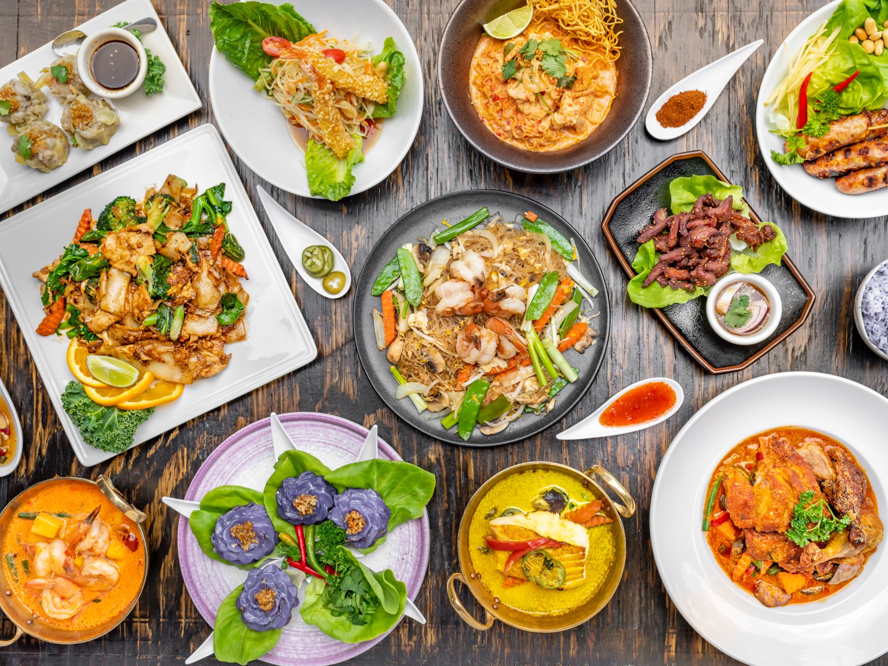 An overhead spread of Nour Thai dishes — curries, pad see ew, glass noodles, dim sum, and som tum — on a rustic wood table